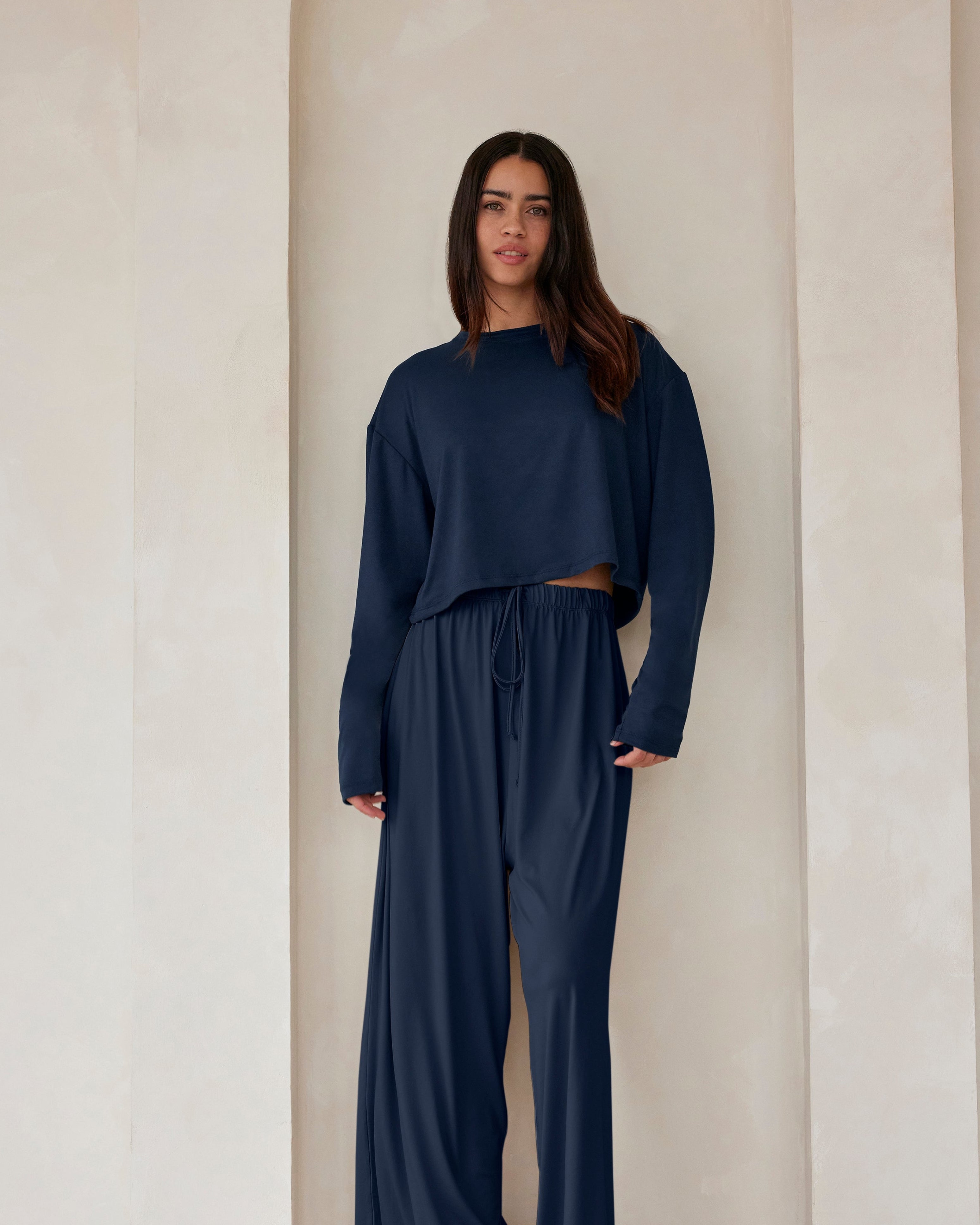 A woman standing against a beige feature wall wearing Bumpsuits Cloud lounge pants and a long sleeve crop tee in navy