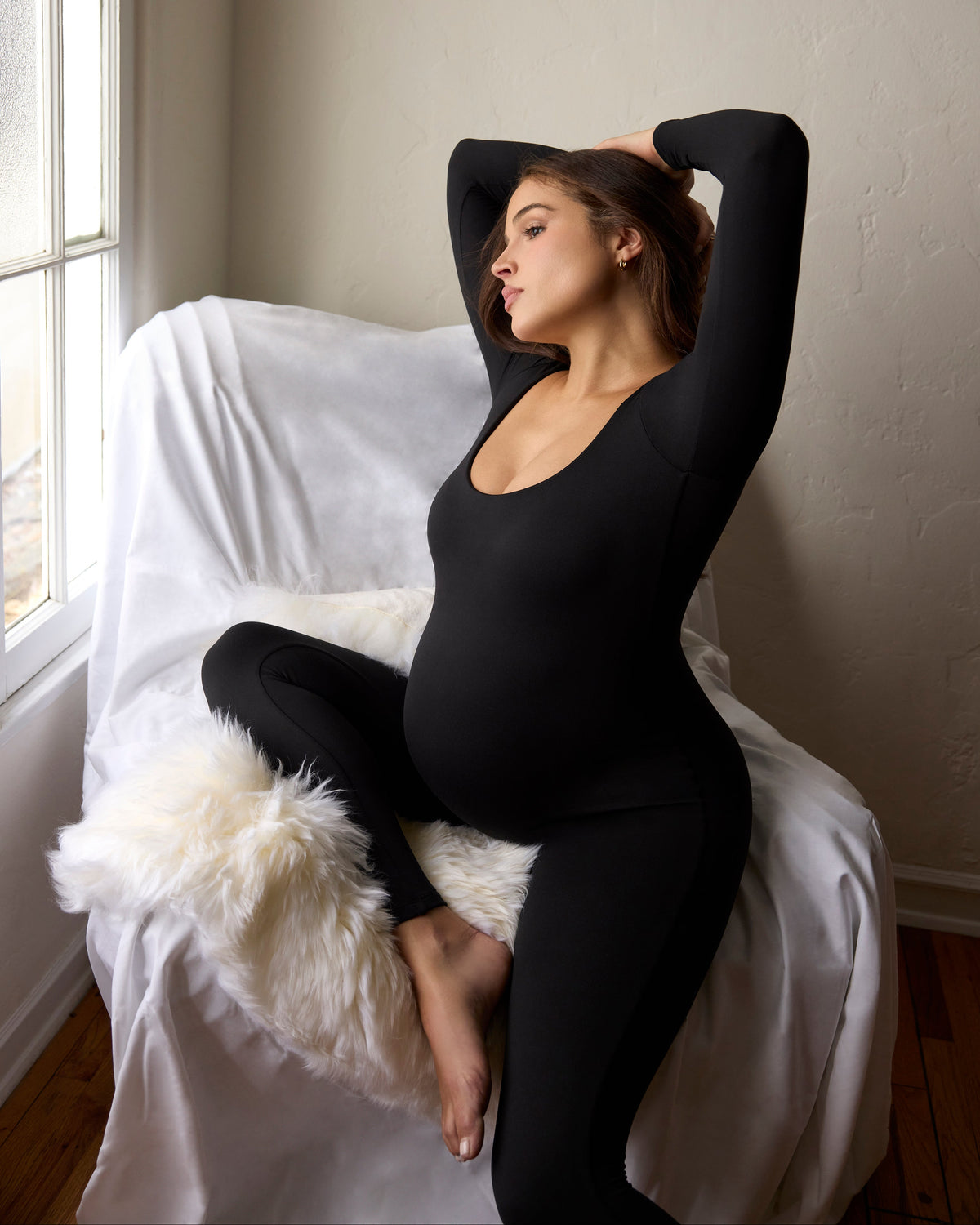 A pregnant woman wearing a black long-sleeve jumpsuit sits on a white-covered chair in a sunlit room with a window, leaning back with her arms raised.