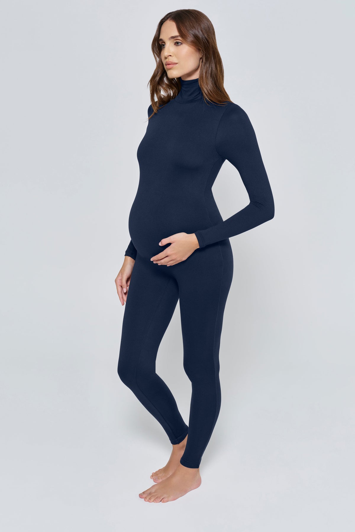 A pregnant woman modeling a navy blue maternity turtleneck jumpsuit against a light gray studio background.