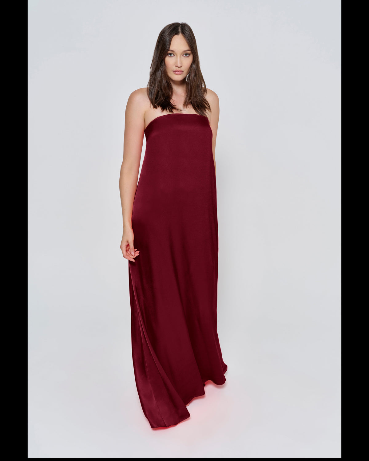 Pregnant woman wearing a Bumpsuit brushed satin gown in a Burgundy color against a white background