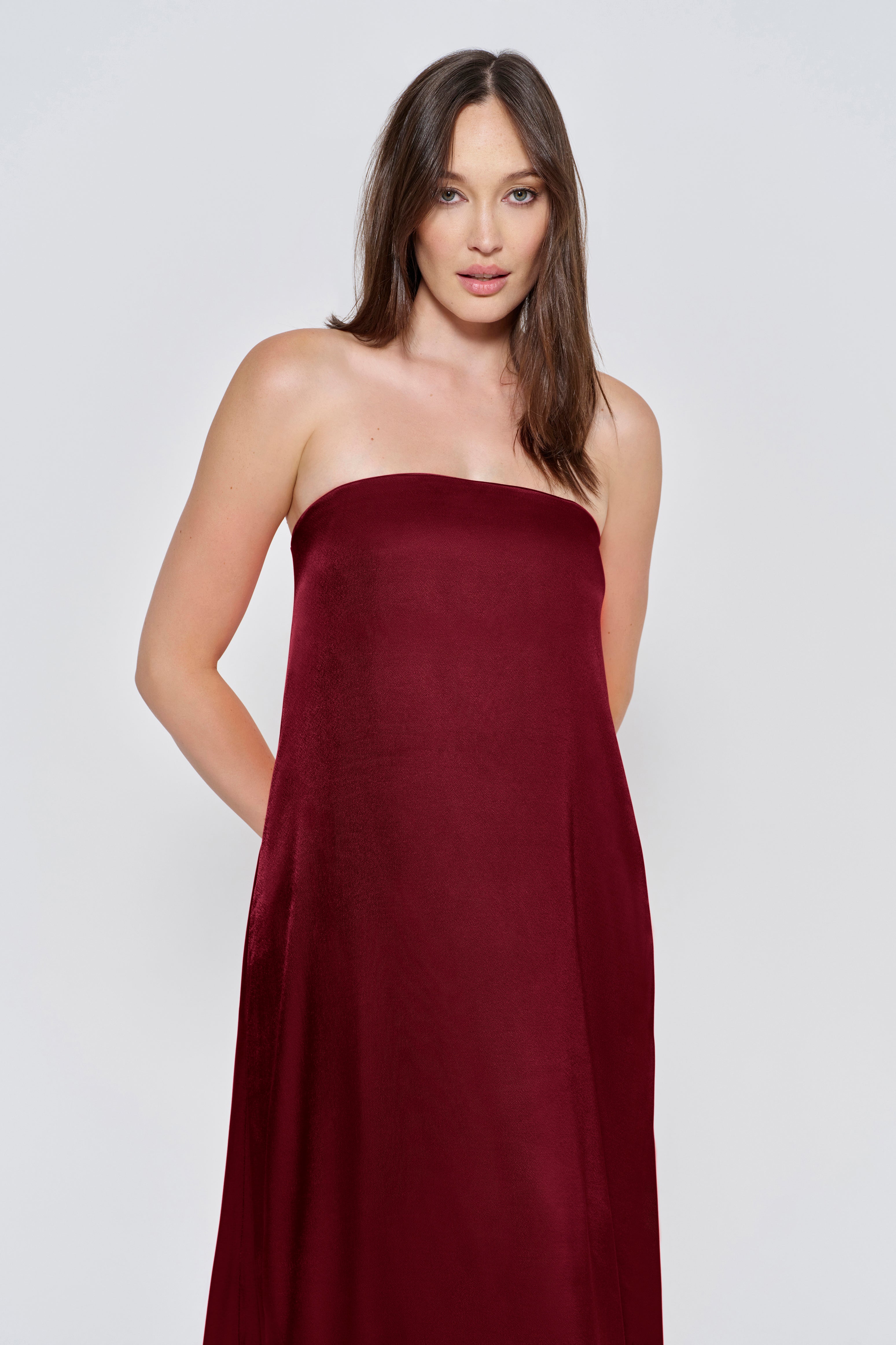 Pregnant woman wearing a Bumpsuit brushed satin gown in a Burgundy color against a white background