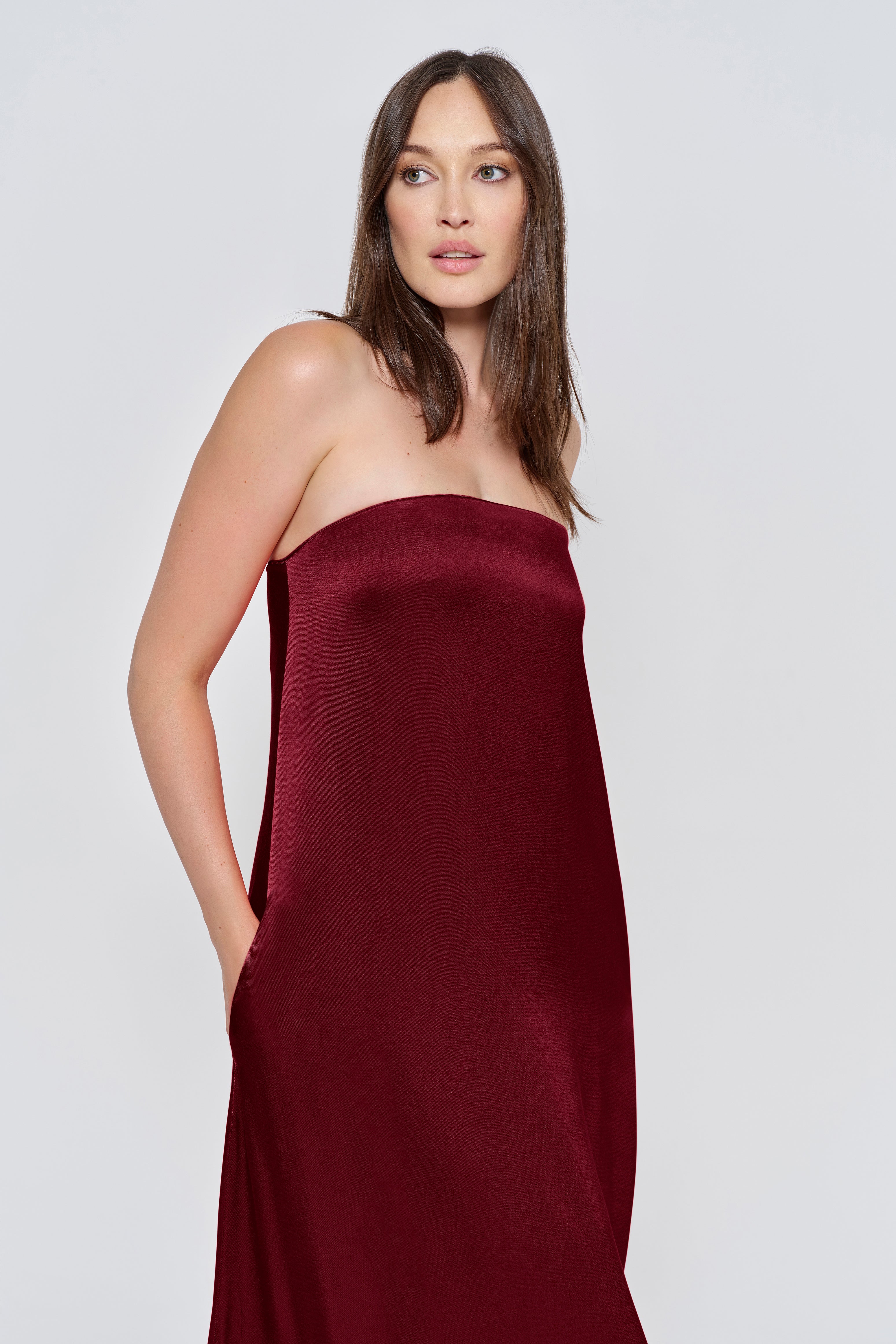 Pregnant woman wearing a Bumpsuit brushed satin gown in a Burgundy color against a white background