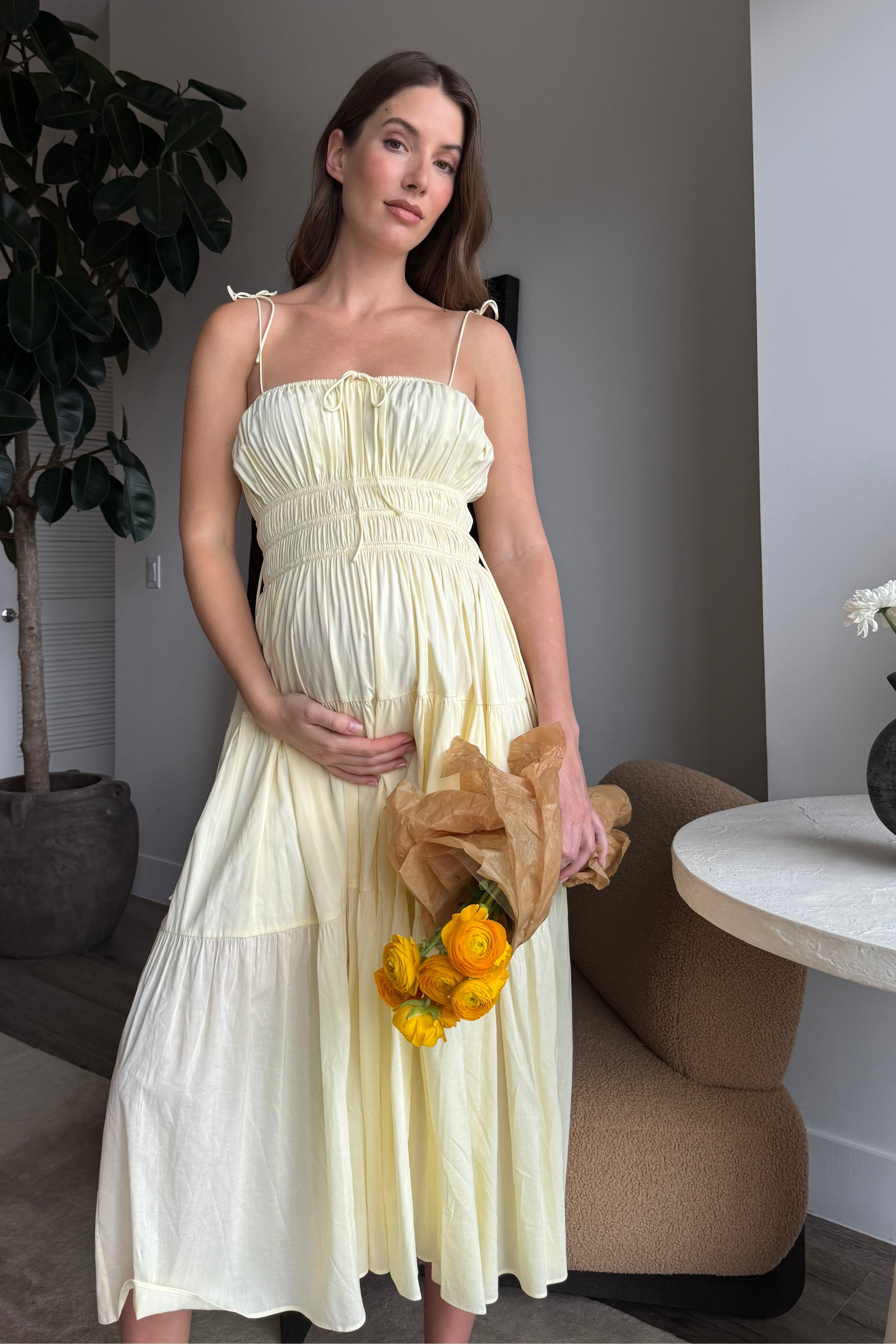 Woman in a light yellow shirred dress holding flowers in a room with a plant and table.