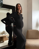 A pregnant woman wearing a fitted black long-sleeve maternity dress stands indoors next to a mirror that reflects her back. The room has white walls, a beige boucle chair, and a side table with a vase.