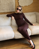 A pregnant woman wearing a soft rib maxi maternity dress in chocolate brown by Bumpsuit, sitting on a beige sofa