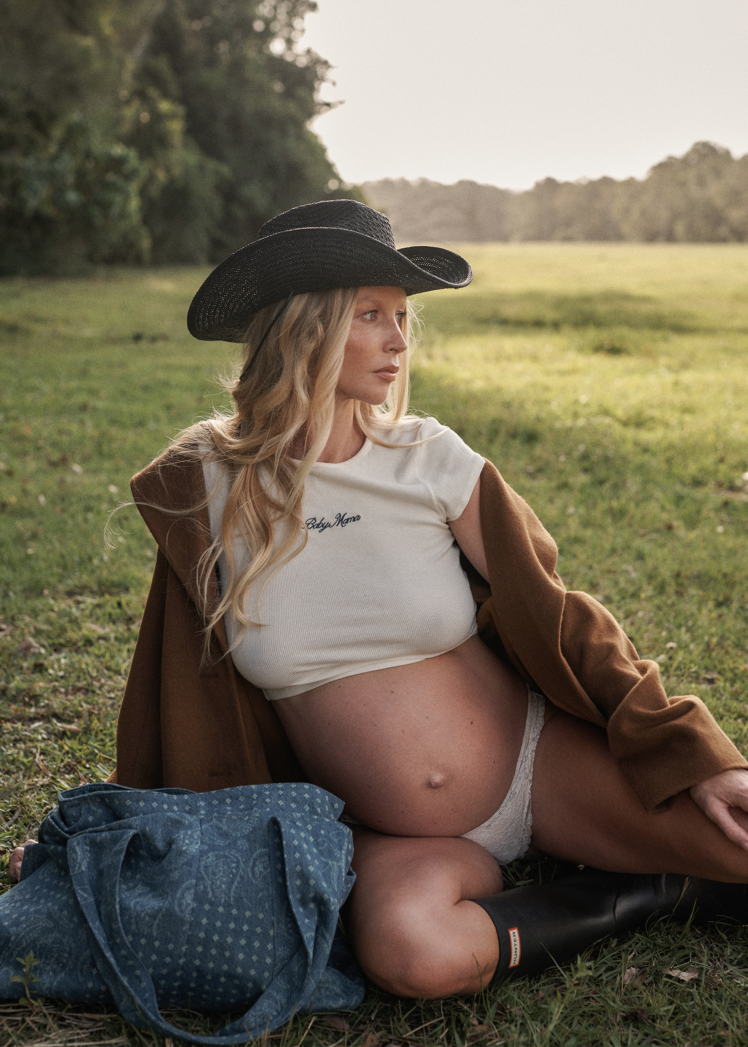 Pregnant woman sitting in a field wearing a wide-brimmed hat and brown coat.