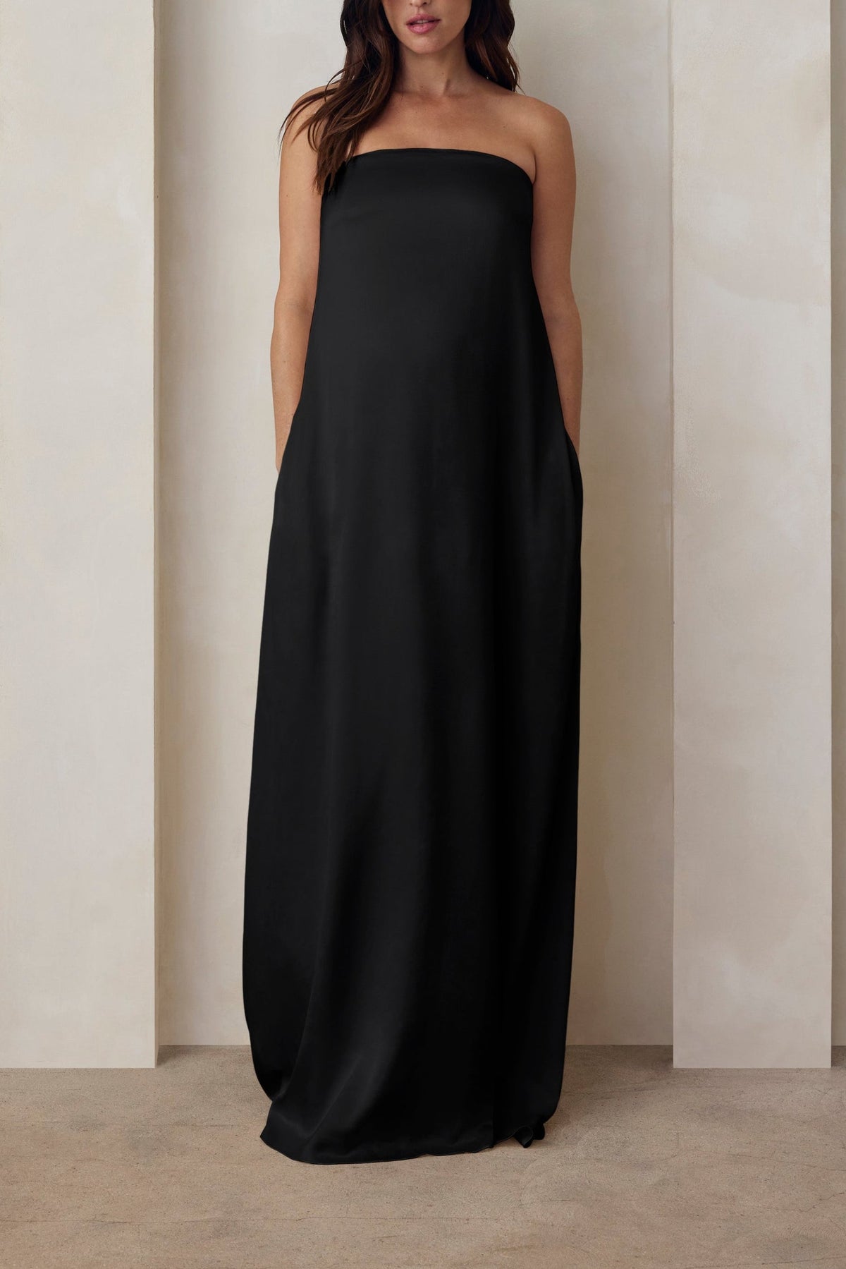 A pregnant woman wearing the Bumpsuit brushed satin strapless maxi maternity gown in black against a beige feature wall