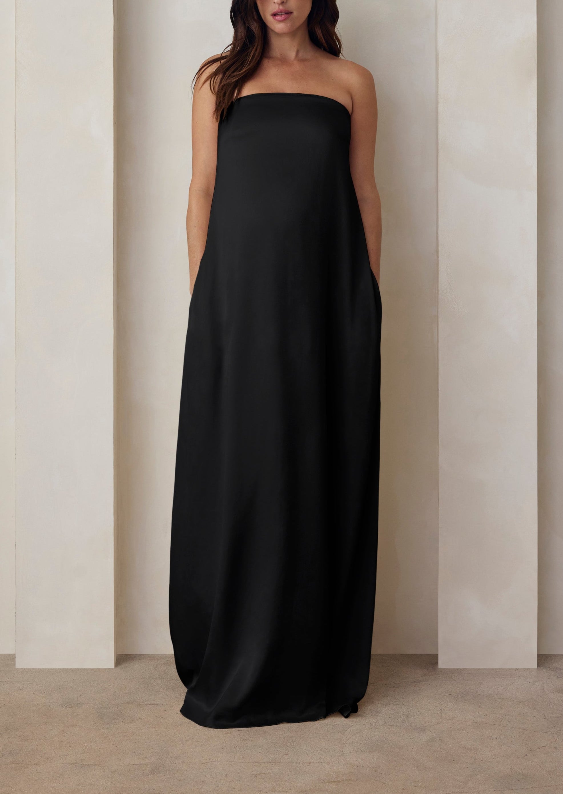 A pregnant woman wearing the Bumpsuit brushed satin strapless maxi maternity gown in black against a beige feature wall