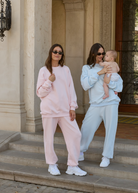 Two individuals, one carrying a baby in a armadillo baby carrier, wearing oversized terry sweatpants in blue and pink colors with white sneakers.