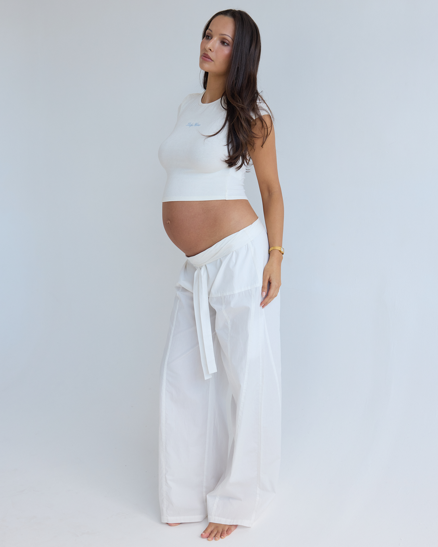 A pregnant woman stands in profile wearing a white sleeveless crop top and matching white wide-leg pants by Bumpsuit, photographed against a plain light gray studio background.