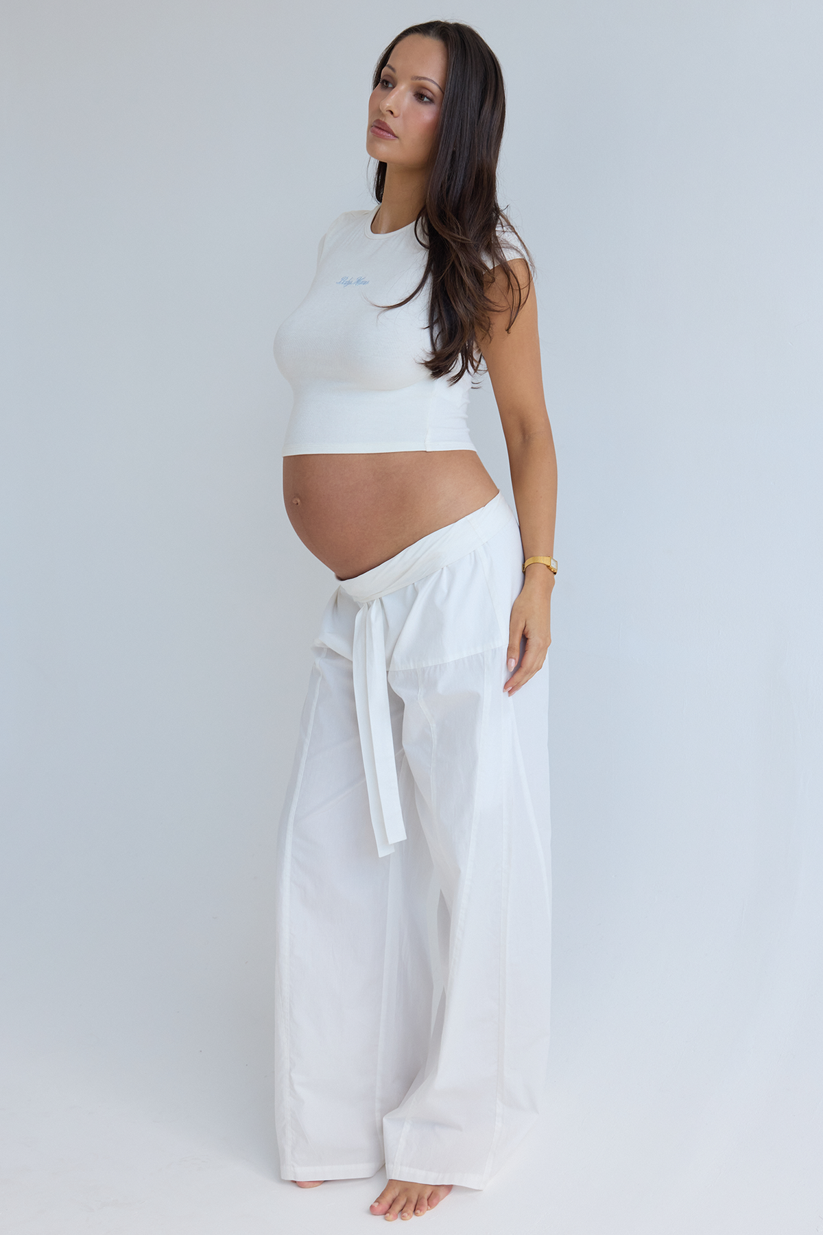Three-quarter view of The Relaxed Pant in white poplin on a pregnant model, showing the foldable waistband and waist tie in a gray studio