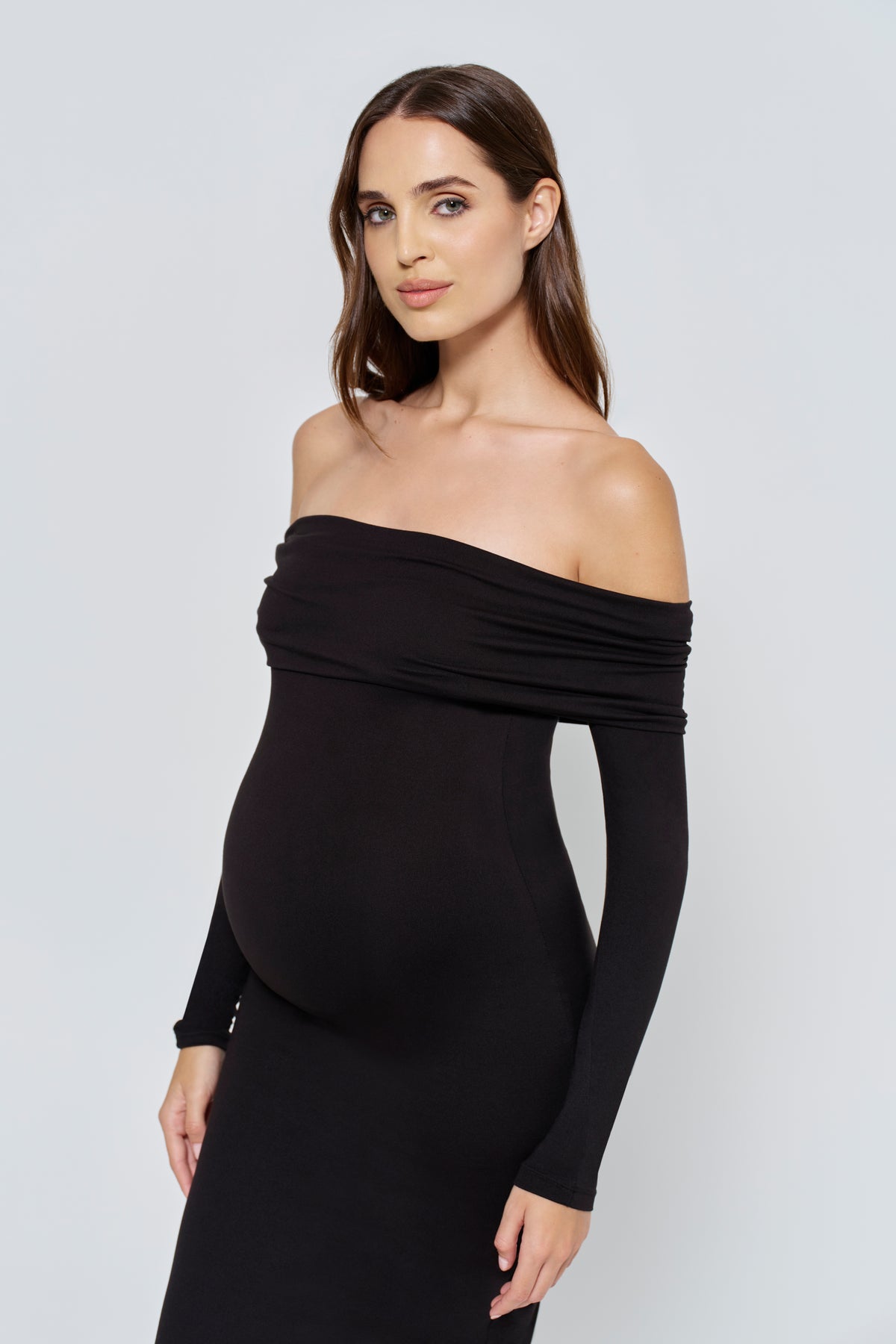 Pregnant woman wearing a black off-shoulder dress with long sleeves against a white background