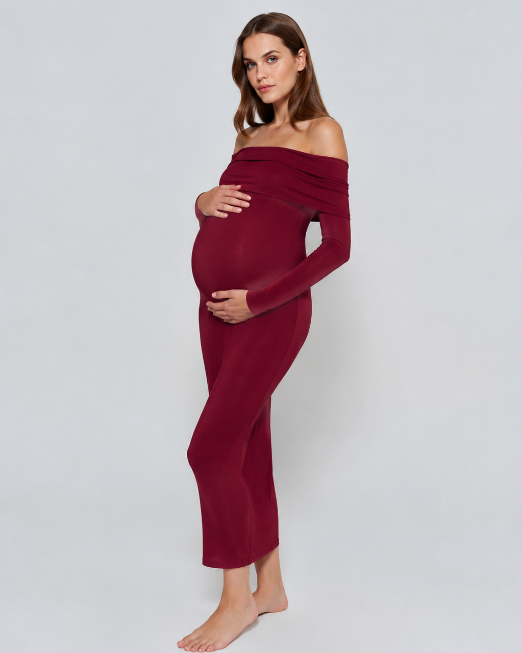 A pregnant woman modeling a long-sleeve, off-the-shoulder maternity dress in deep burgundy against a light gray studio background.