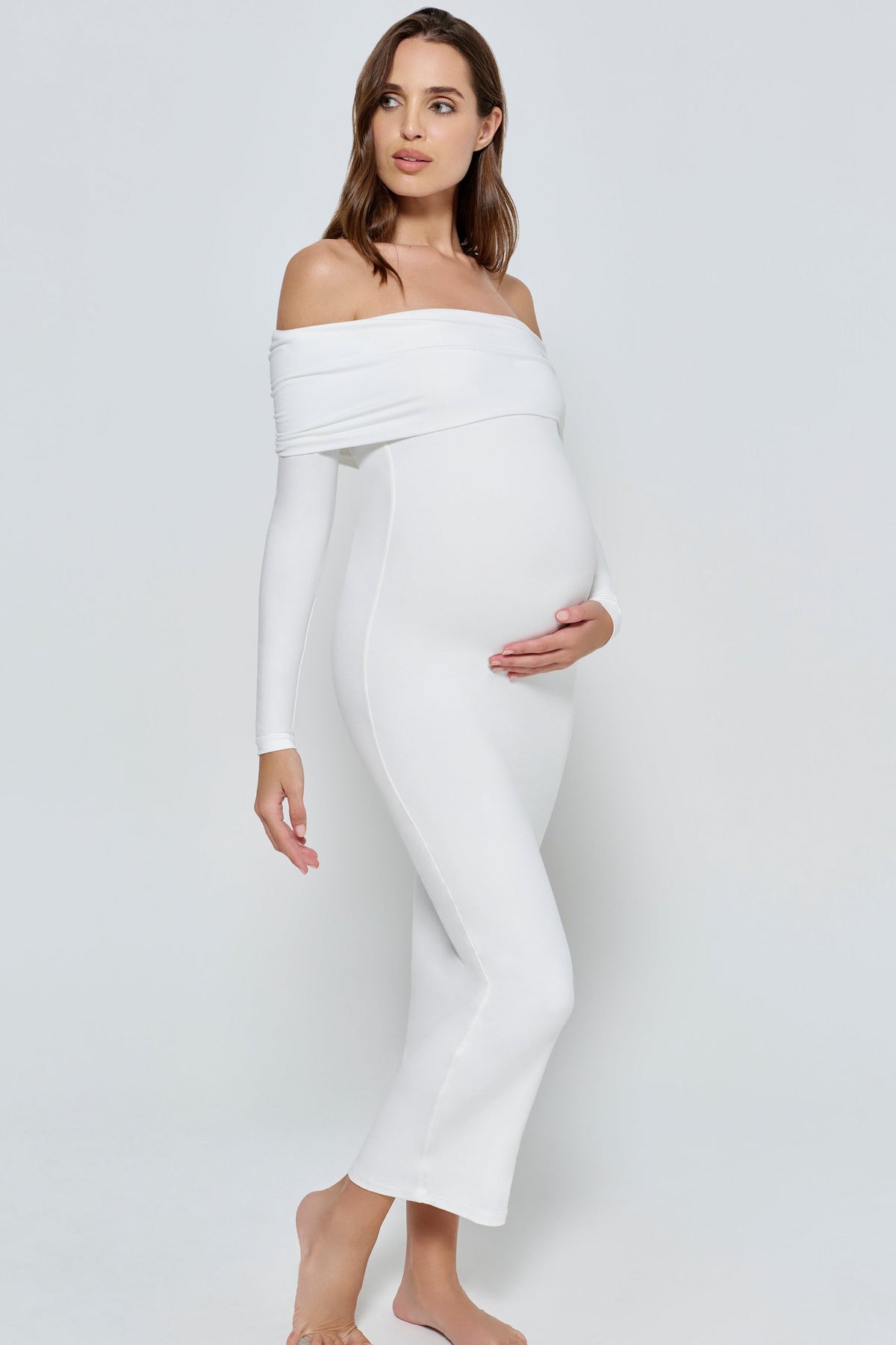 Pregnant woman wearing a ivory Bumpsuit off-shoulder dress on a white background