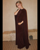 A pregnant woman wearing a Bumpsuit maxi dress with a floor length cape detail in chocolate brown against a neutral setting