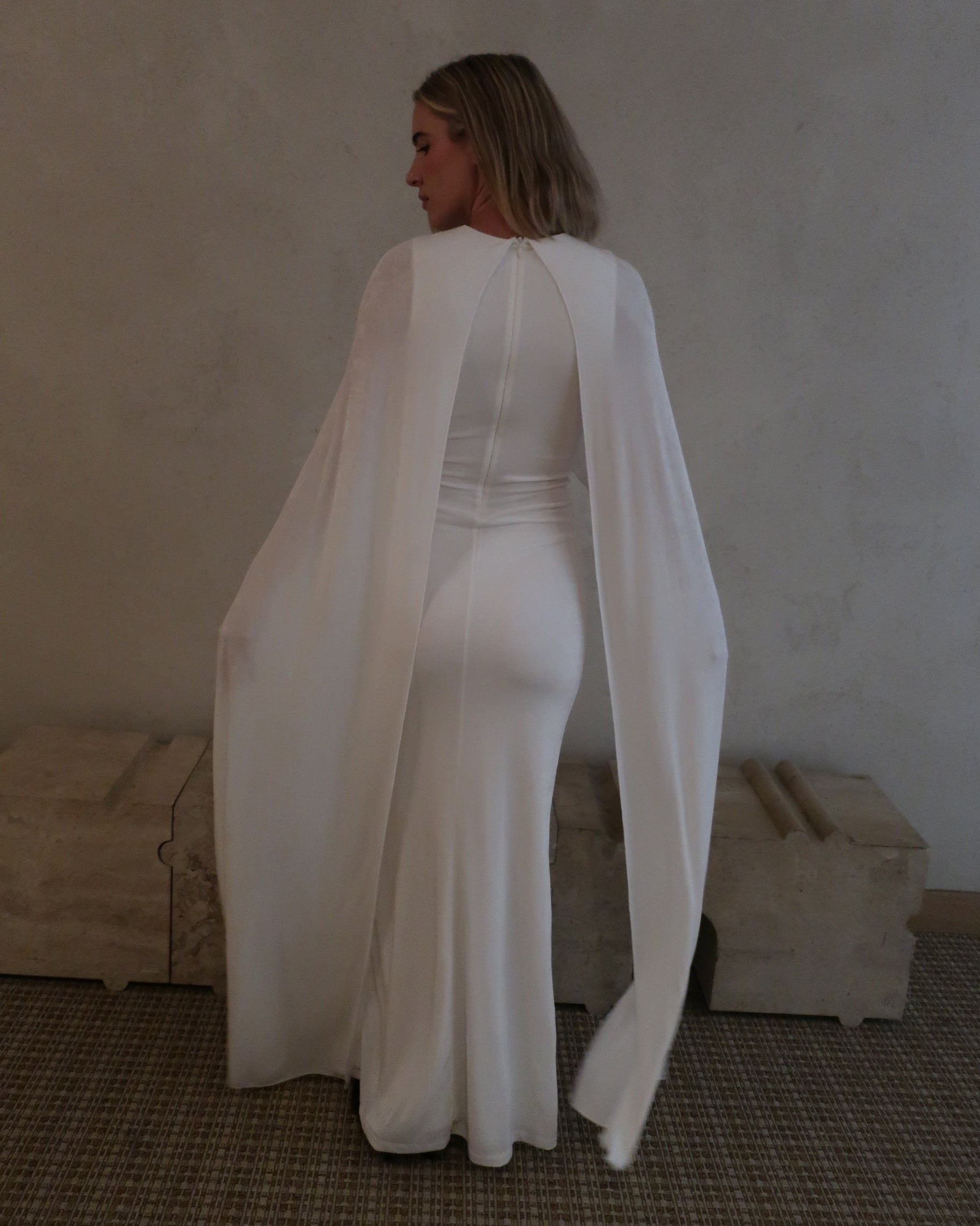 A woman wearing Bumpsuit maxi gown with a long sheer cape, standing in a neutral indoor setting.