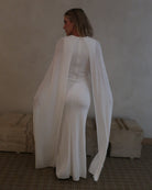 A woman wearing Bumpsuit maxi gown with a long sheer cape, standing in a neutral indoor setting.