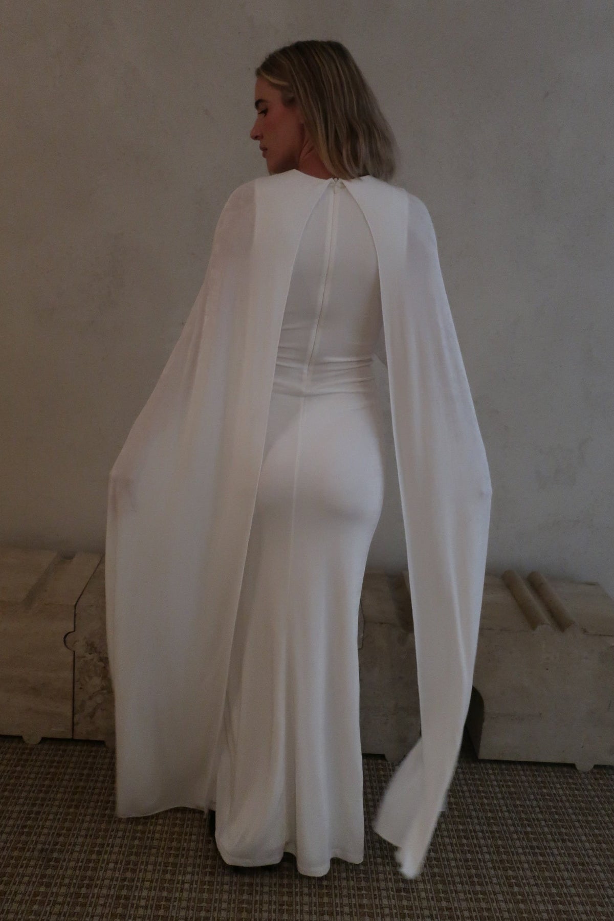 Back view of a woman wearing Bumpsuit maxi gown with a long sheer cape, standing in a neutral indoor setting.