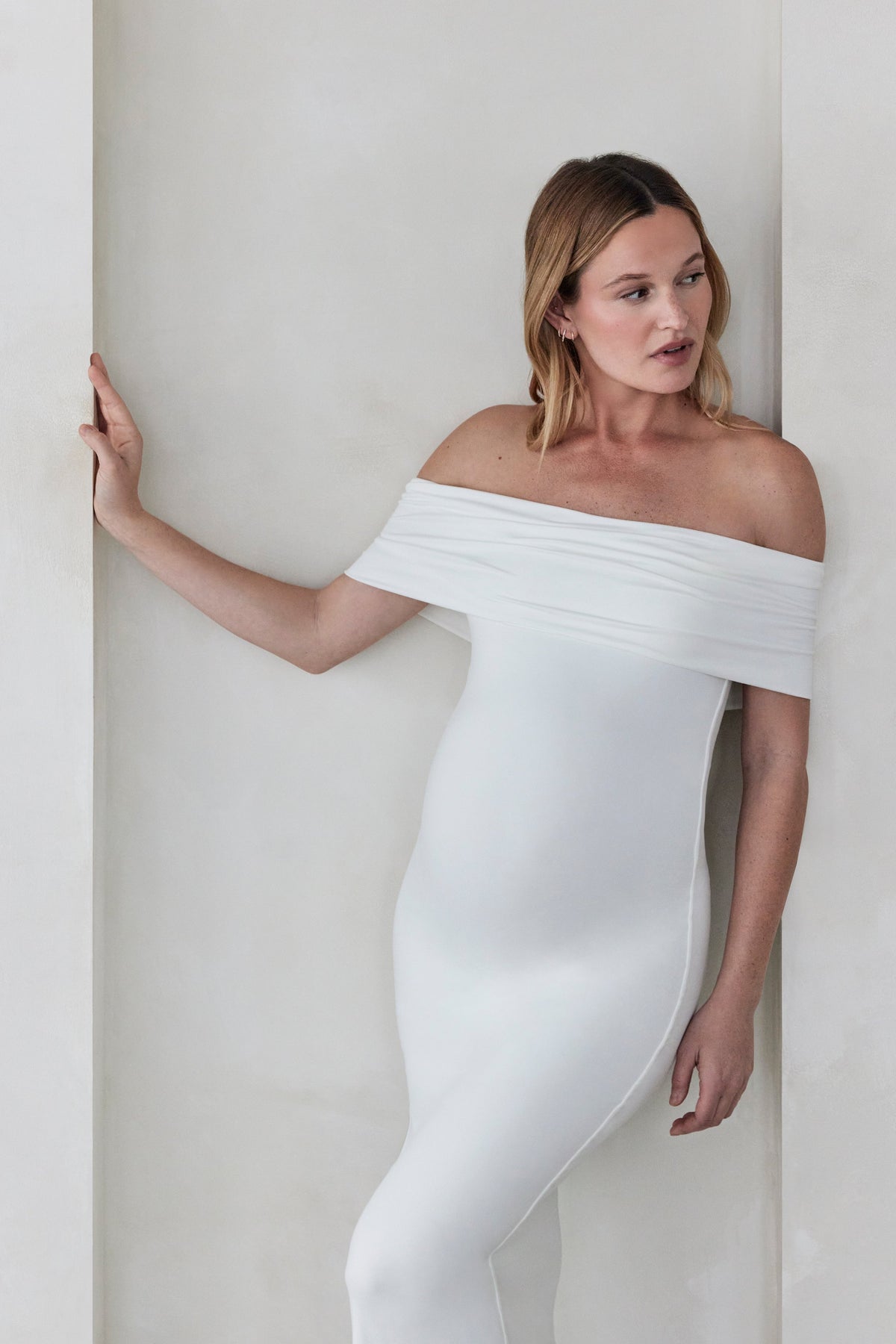 Pregnant woman wearing Bumpsuits off the shoulder Ivory Bianca maternity maxi dress standing  against a beige wall