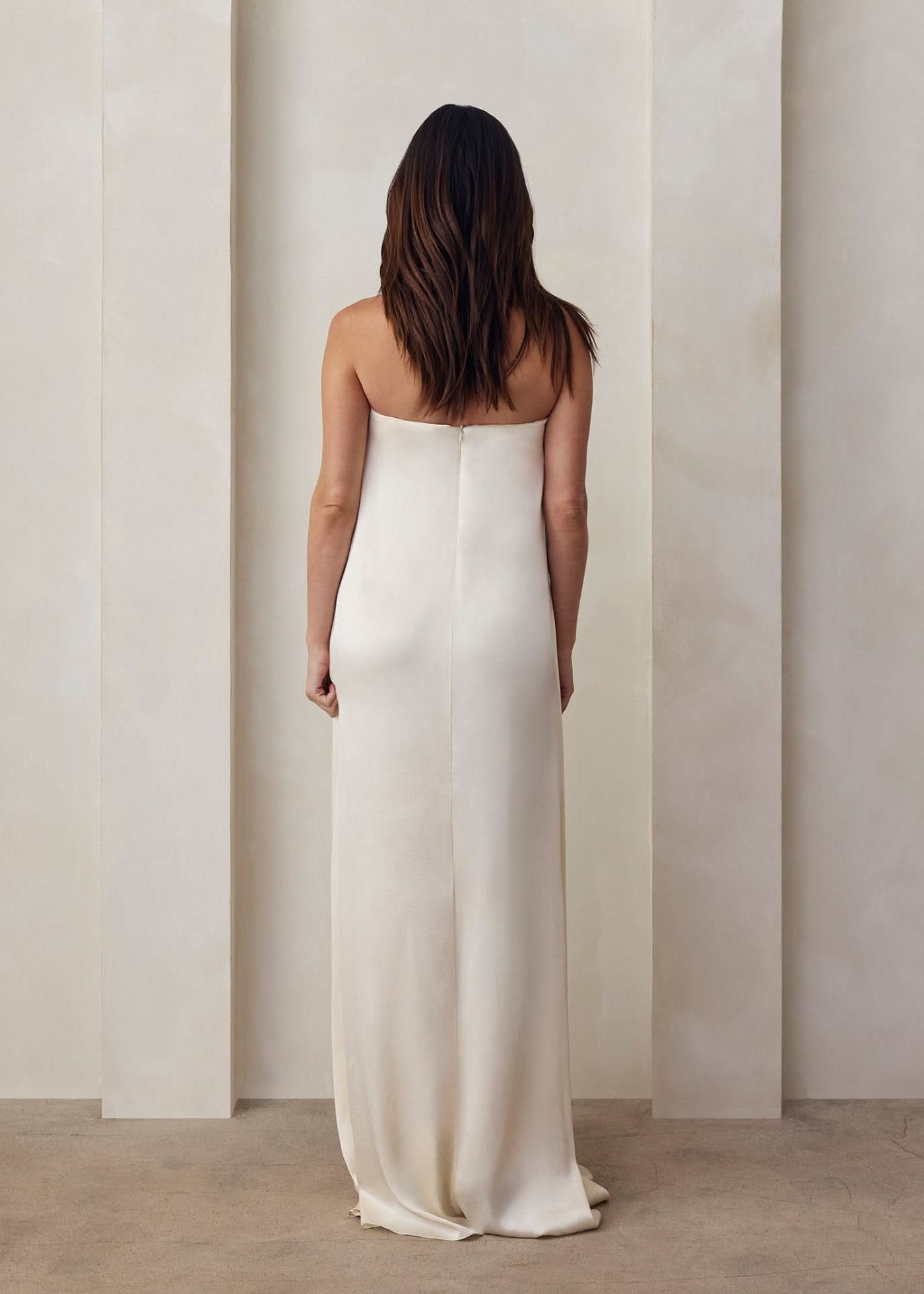 the back view of a pregnant model wearing the Bumpsuit brushed satin gown in ivory against a beige feature wall
