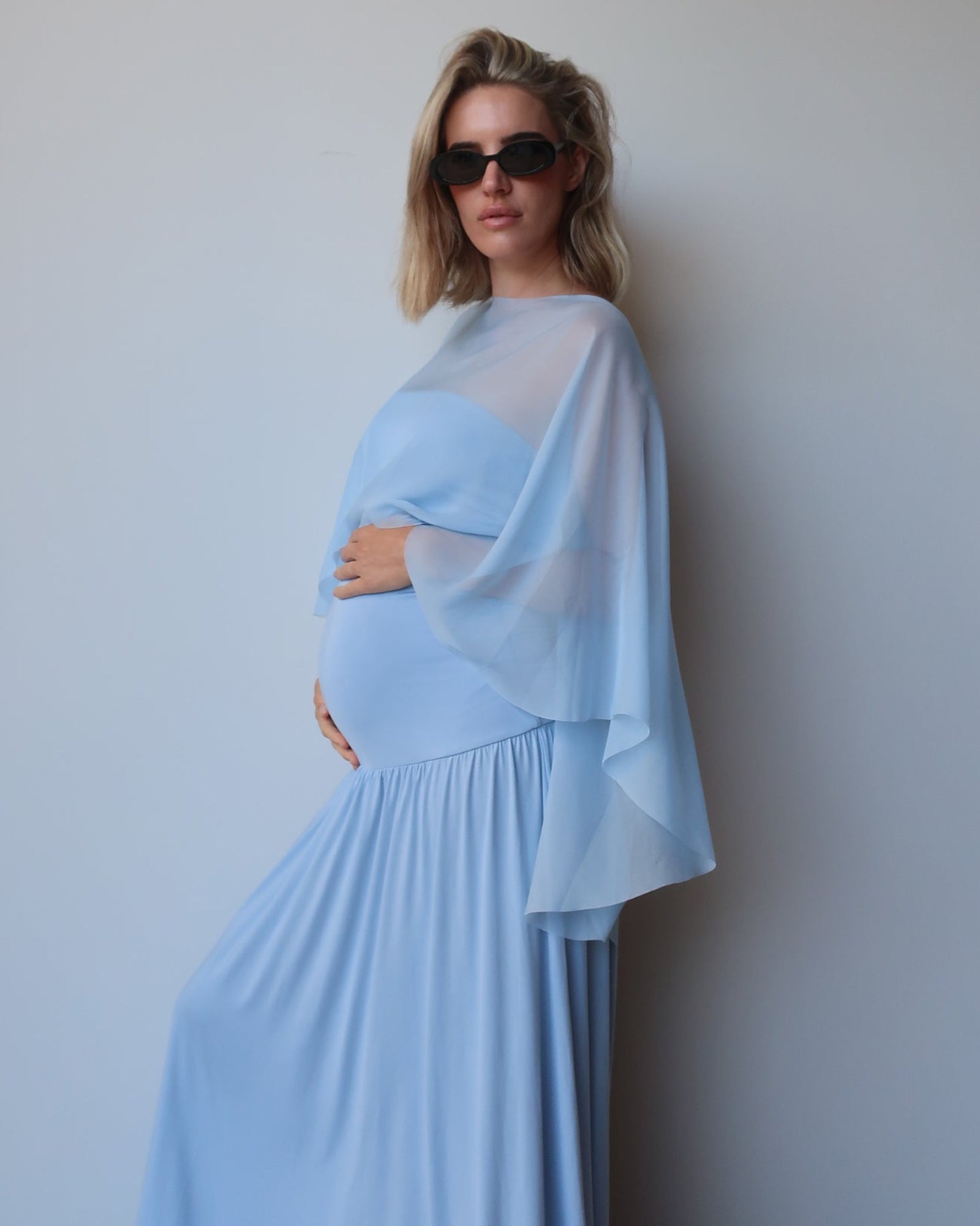 A pregnant woman wearing a light blue maternity dress with a sheer chiffon cape by bumpsuit and sunglasses, posing against a plain light gray wall.