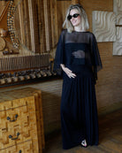 A woman wearing Bumpsuits Maria dress and chiffon Capelet in black, styled with black sunglasses, standing in a rustic room with wooden furniture and wall art.