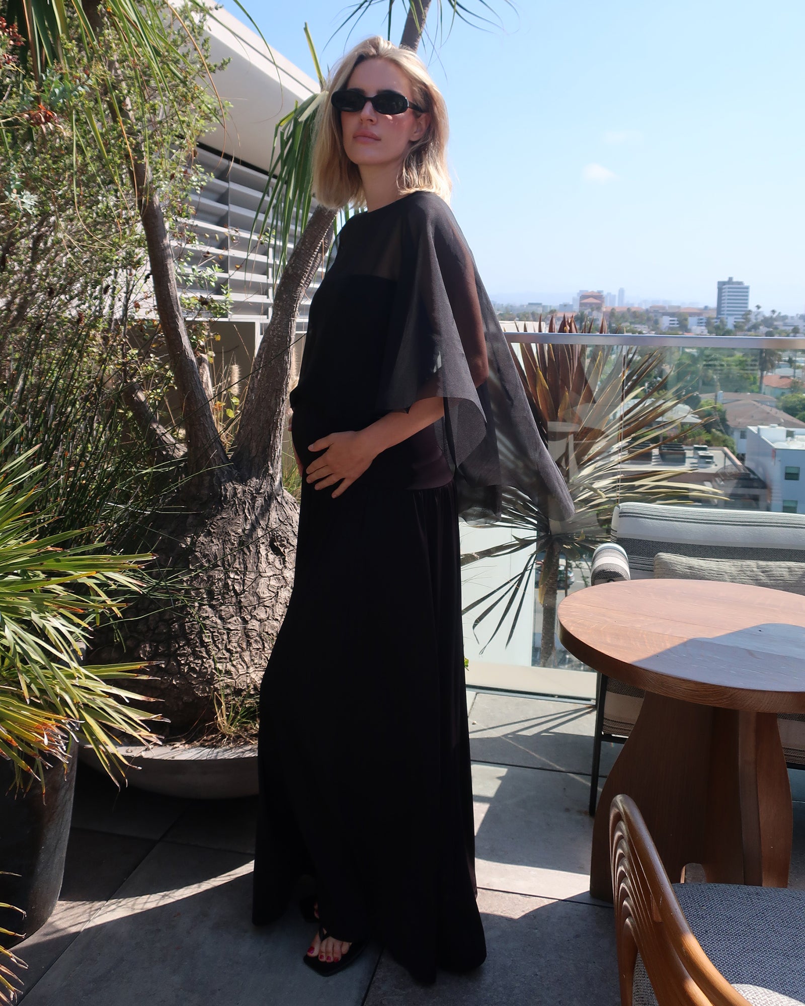 A woman wearing the black Bumpsuit Maria maxi dress with sheer chiffon cape  and black sunglasses, standing on a sunny outdoor balcony with plants and a table in the background.