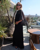 A woman wearing the black Bumpsuit Maria maxi dress with sheer chiffon cape  and black sunglasses, standing on a sunny outdoor balcony with plants and a table in the background.
