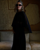 A woman wearing a Bumpsuit long black dress with a chiffon capulet and black sunglasses, standing in a dimly lit room with a textured beige sofa and wall art behind her.