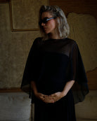 A woman with blonde hair wearing a black sheer cape-sleeve and dress by Bumpsuit, styled with black sunglasses, standing in a dimly lit room.