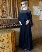 A pregnant woman wearing a long black maternity dress by Bumpsuit with a chiffon capulet and black sunglasses, standing in a modern room with wood-paneled walls and decorative wooden furniture.
