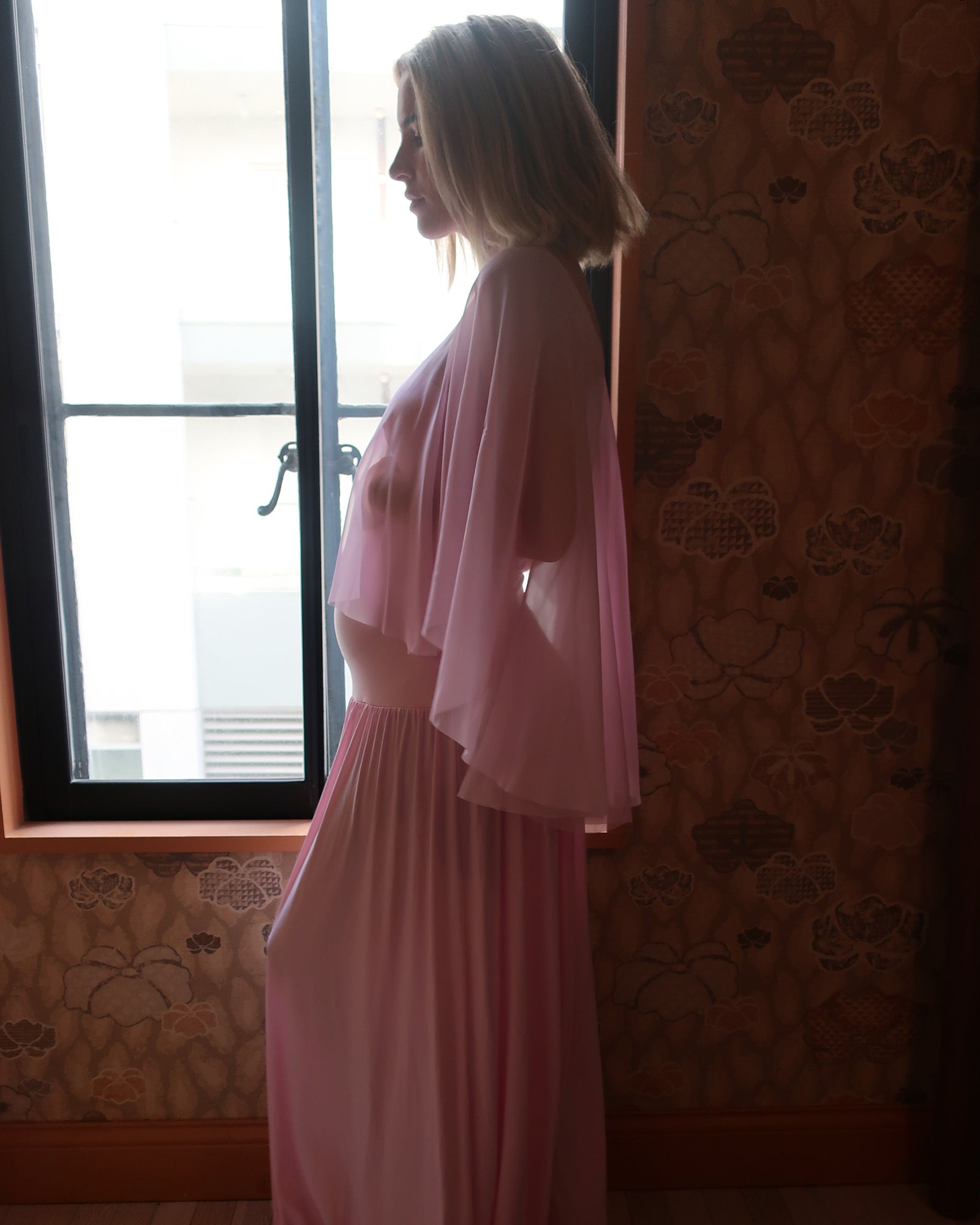A pregnant woman in profile wearing a long pink dress by Bumpsuit with a sheer chiffon cape, standing indoors near a window with patterned wallpaper.