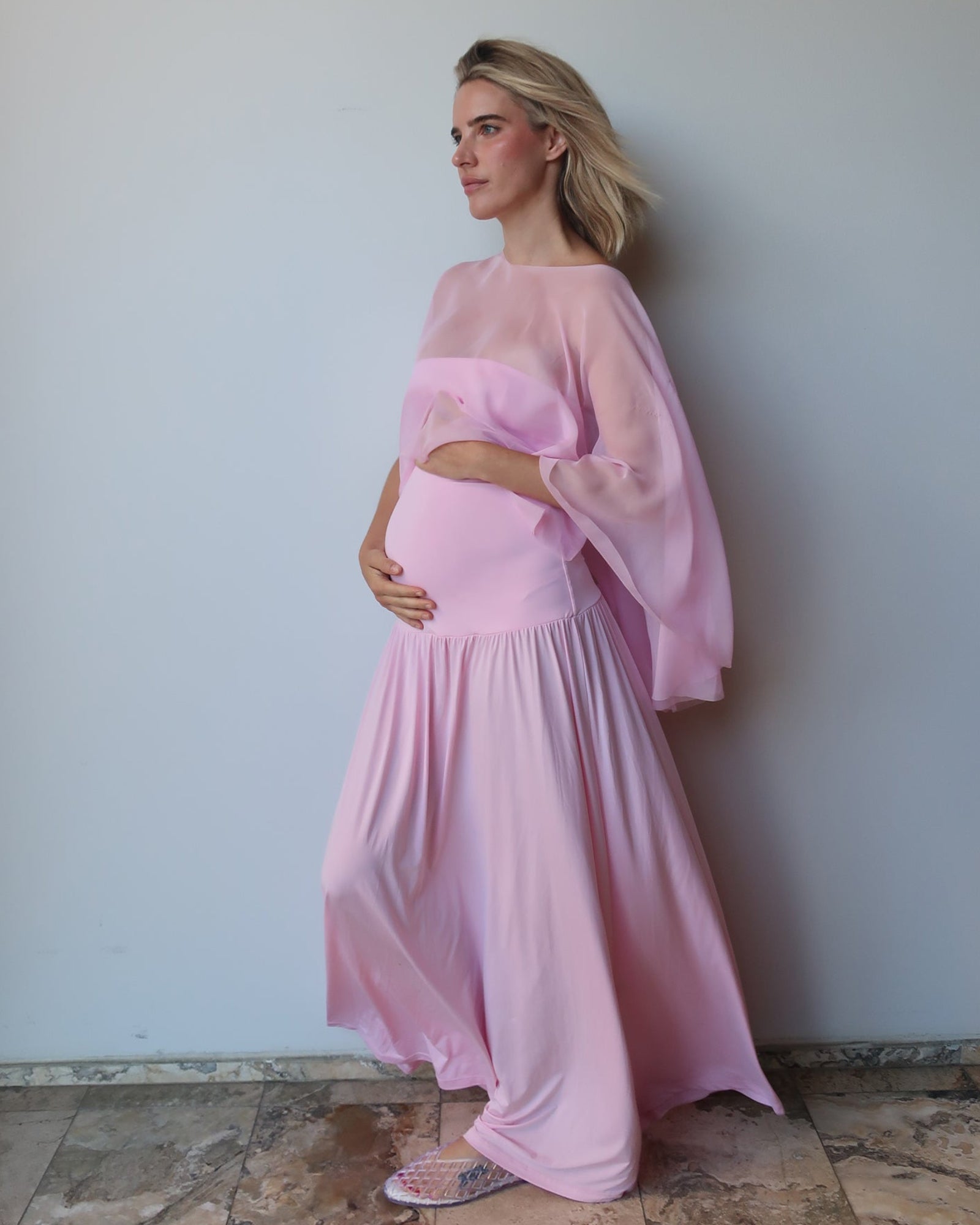A pregnant woman wearing a light pink maternity dress with a sheer chiffon cape, standing against a plain light gray wall on a stone-tiled floor.