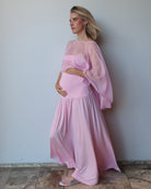 A pregnant woman wearing a light pink maternity dress with a sheer chiffon cape, standing against a plain light gray wall on a stone-tiled floor.