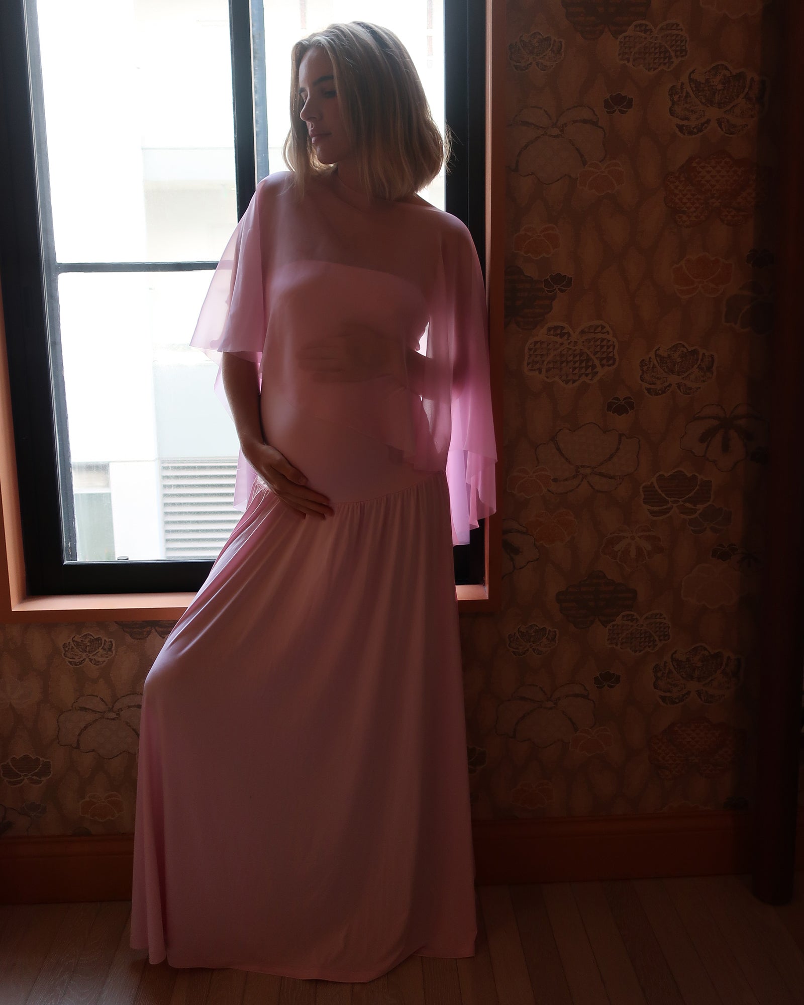 A pregnant woman wearing a Bumpsuit long, flowing light pink gown with a chiffon cape  stands in front of a window, with patterned wallpaper and wood flooring visible in the room.