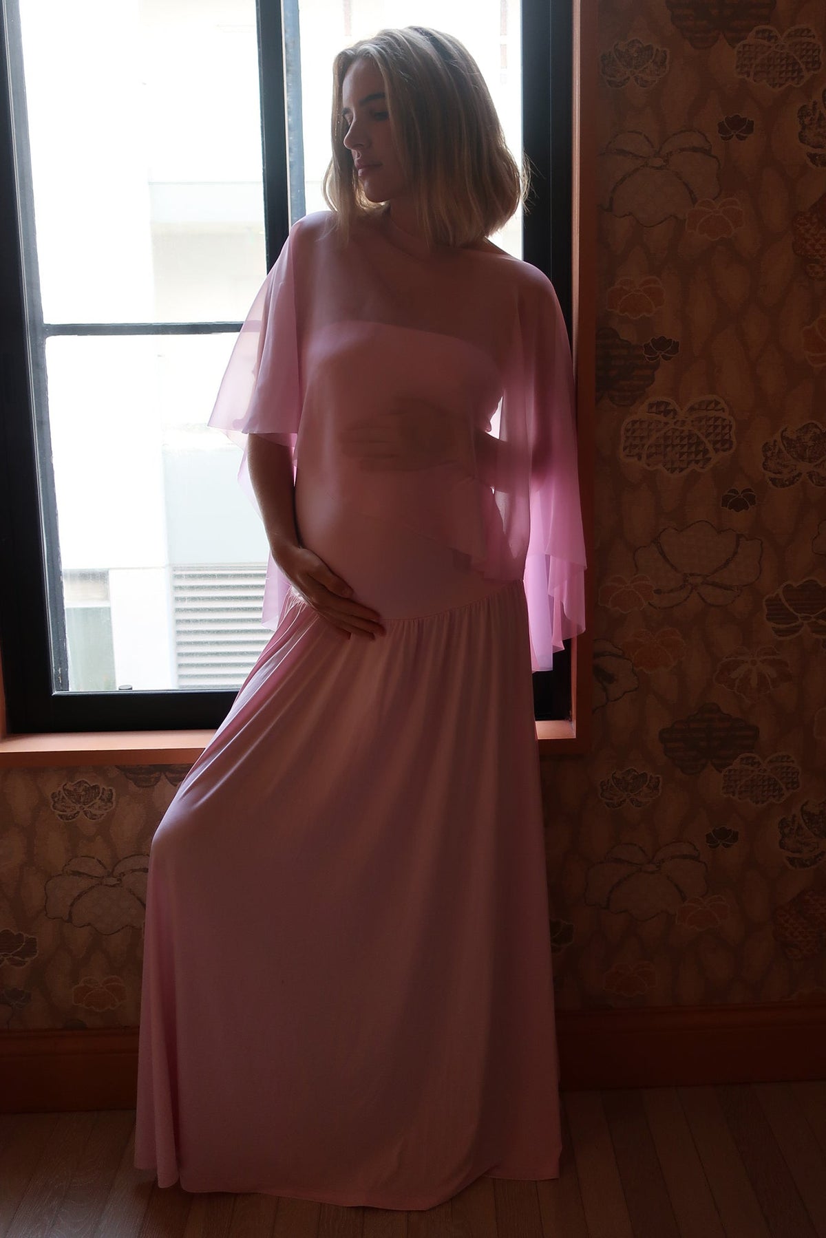Full-length view of The Chiffon Capelet showcasing its sheer pink chiffon drape on a pregnant model by a window