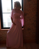 A pregnant woman wearing a Bumpsuit long, flowing light pink gown with a chiffon cape  stands in front of a window, with patterned wallpaper and wood flooring visible in the room.