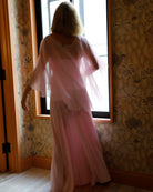 A woman viewed from behind wearing a light pink dress and chiffon cape by Bumpsuit, standing in front of a window in a room with patterned wallpaper.