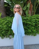 A pregnant woman wearing a Bumpsuit light blue maxi dress with a chiffon cape and sunglasses stands outdoors in front of a white wall and green foliage.