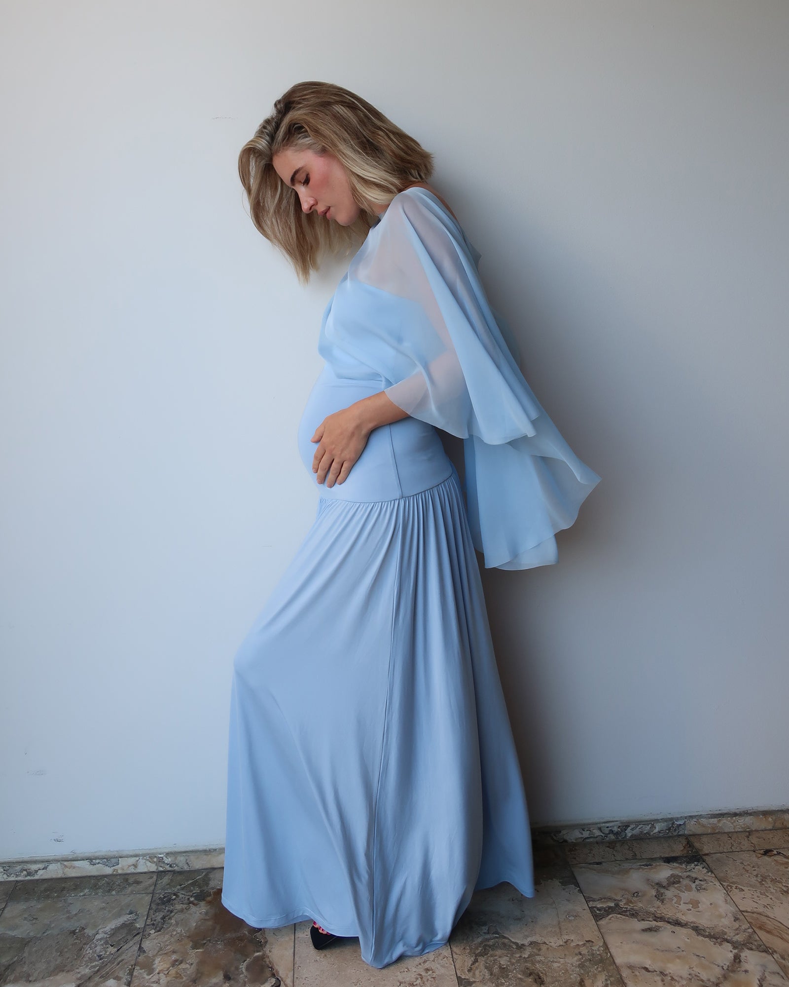 A pregnant woman wearing a light blue maternity maxi dress with a sheer chiffon cape, posing against a plain light gray wall.
