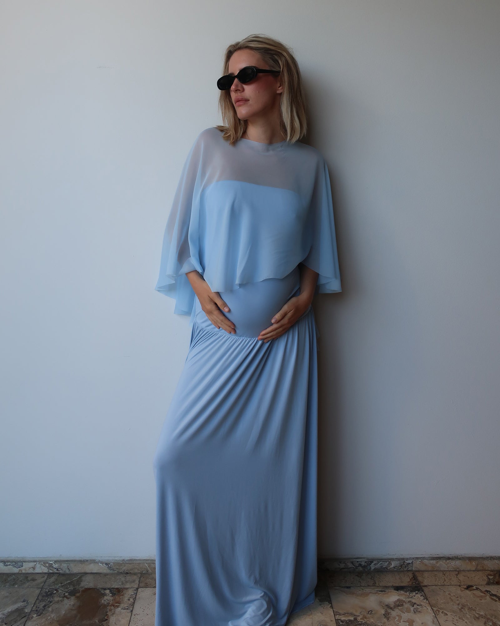 A pregnant woman wearing a  Bumpsuit light blue maternity dress with a chiffon cape, standing against a plain light gray wall.