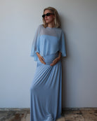 A pregnant woman wearing a  Bumpsuit light blue maternity dress with a chiffon cape, standing against a plain light gray wall.