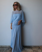 A pregnant woman wearing a light blue maxi dress with a chiffon cape, standing against a plain light gray wall.