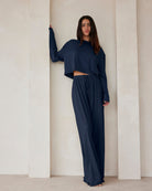 A woman standing against a beige wall in navy lounge pants and a crop long sleeve tee by Bumpsuit