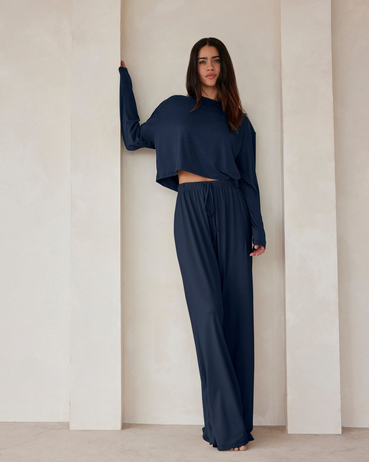 A woman standing against a beige wall in navy lounge pants and a crop long sleeve tee by Bumpsuit