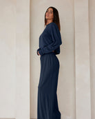 A woman standing against a beige feature wall wearing Bumpsuits Cloud lounge pants and a long sleeve crop tee in navy