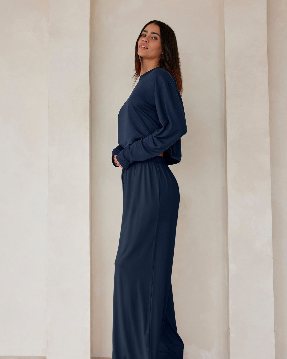 A woman standing against a beige feature wall wearing Bumpsuits Cloud lounge pants and a long sleeve crop tee in navy