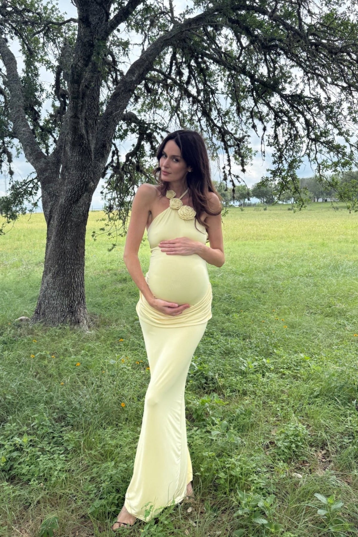 Pregnant woman in a yellow  bumsuit maternity halter gown standing in a grassy field with a tree