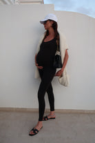 Pregnant woman in black outfit with white cap sitting on a white chair against a light grey wall.