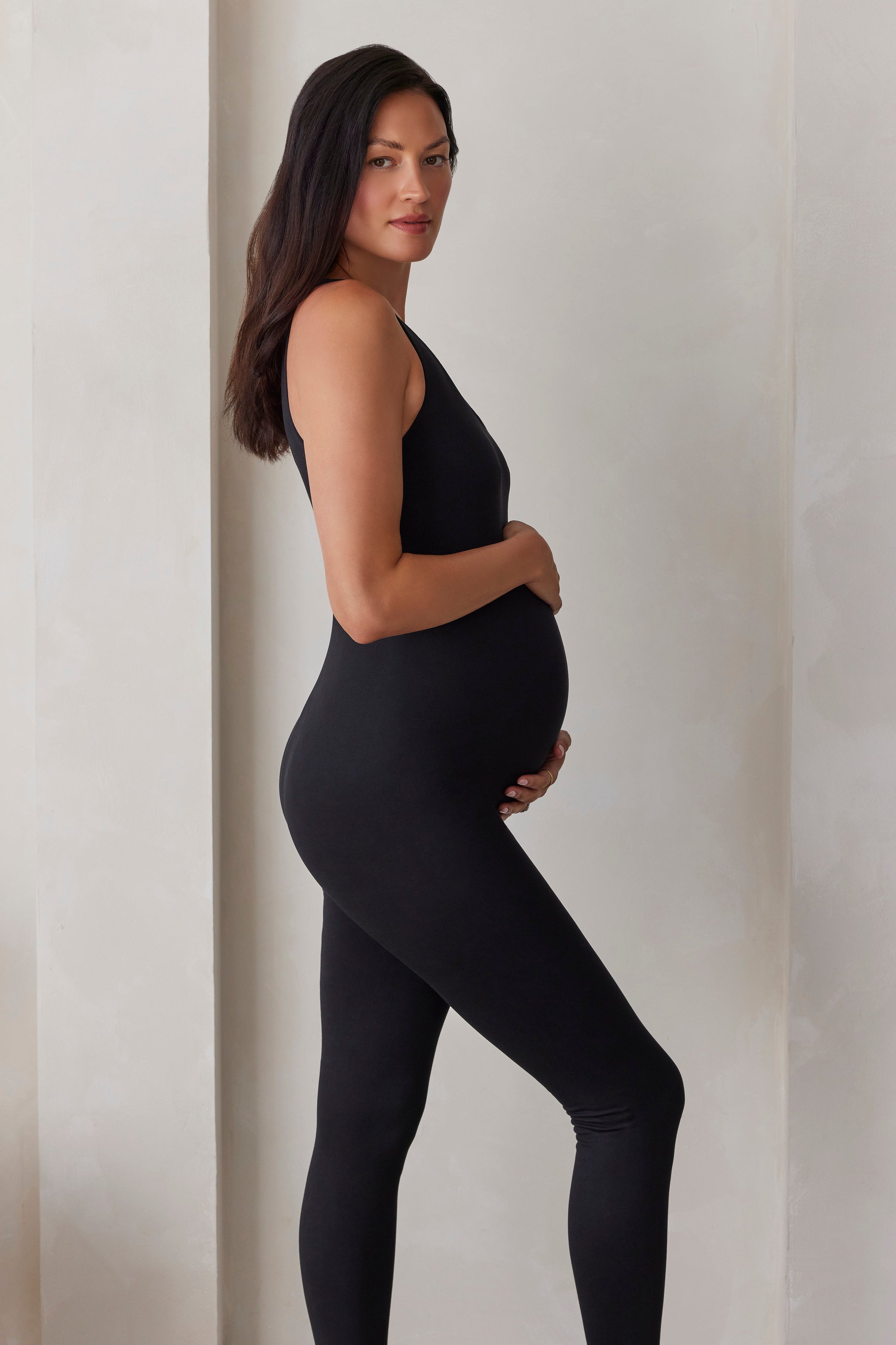 A pregnant woman wearing a black jumpsuit, standing in profile against a neutral beige wall.